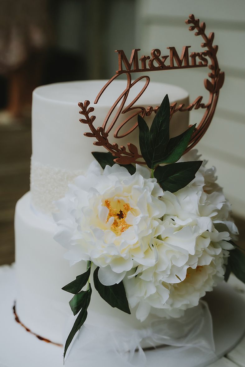 A white wedding cake topper reading 'Mr & Mrs' with delicate white peony flowers and green leaves as decoration.