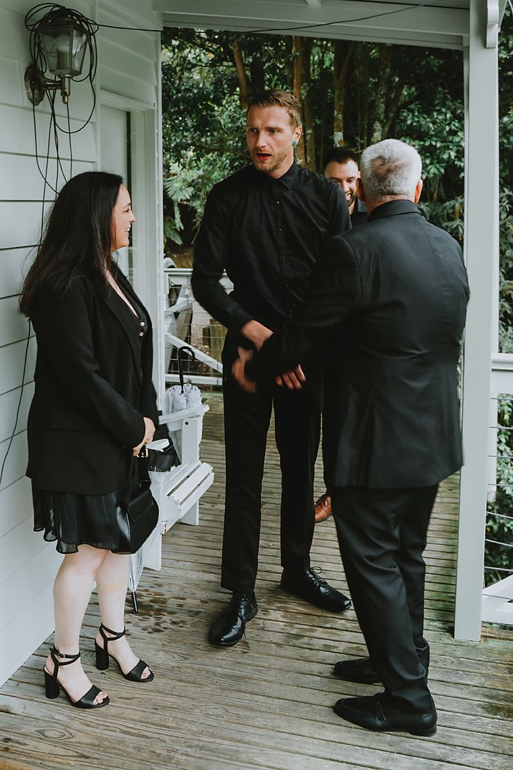 People in black formal attire gather on a wooden porch during what appears to be a social event.