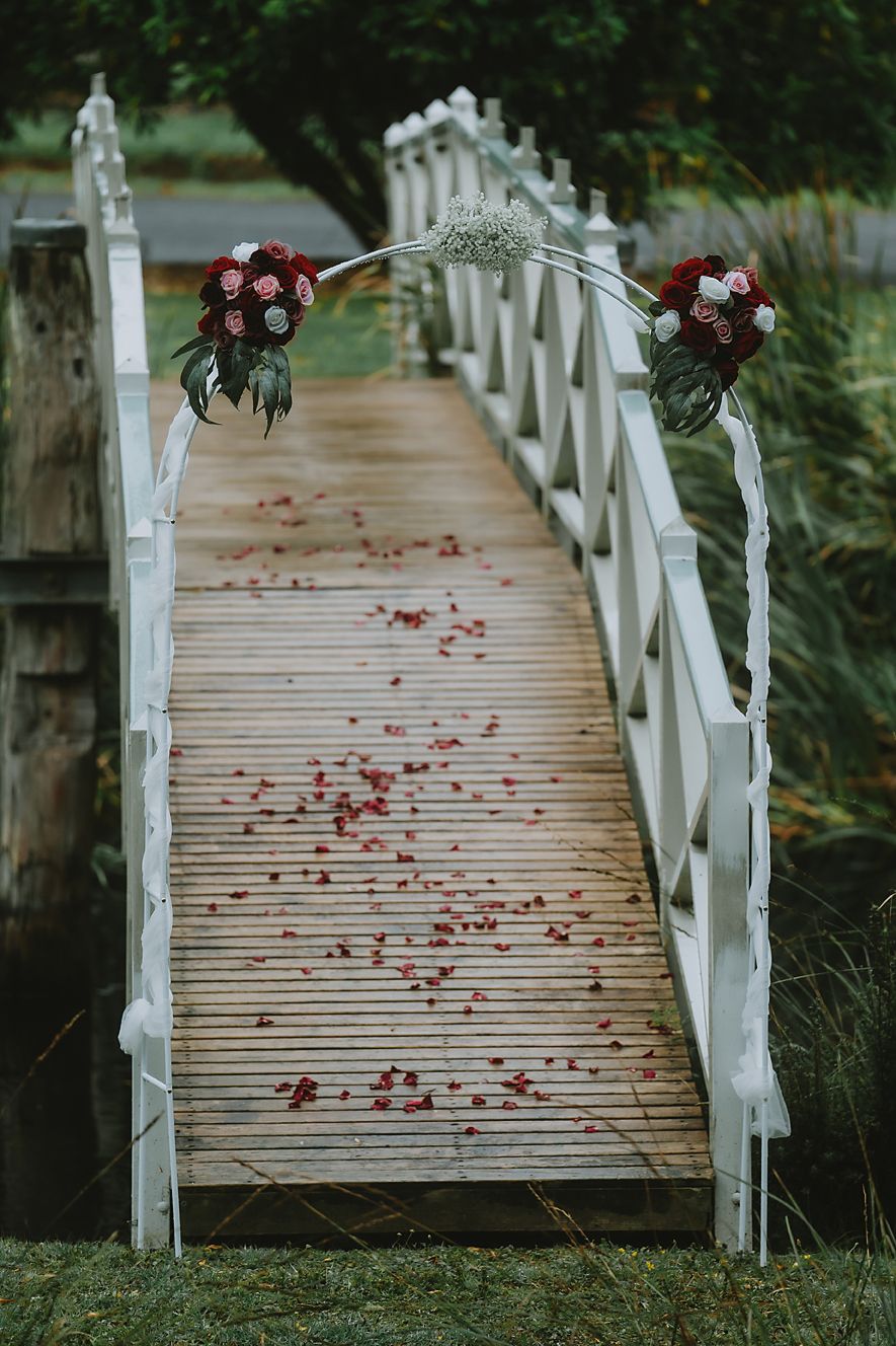 A white wooden footbridge decorated with red roses and scattered petals creates a romantic garden pathway.