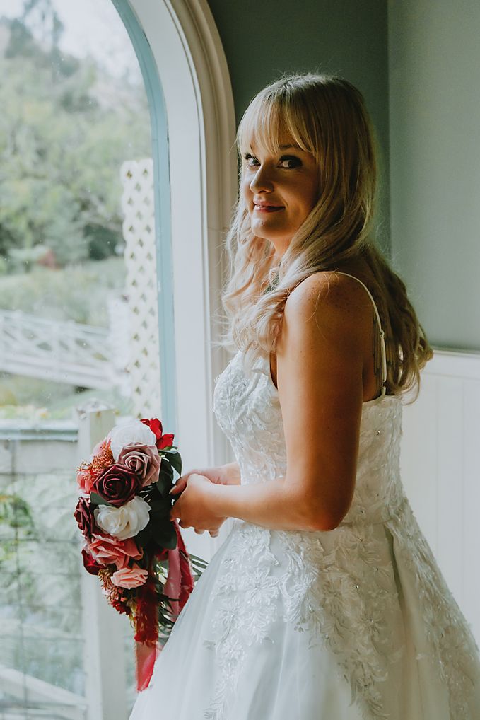 A bride in a white lace dress poses by a window while holding a red and white bouquet.