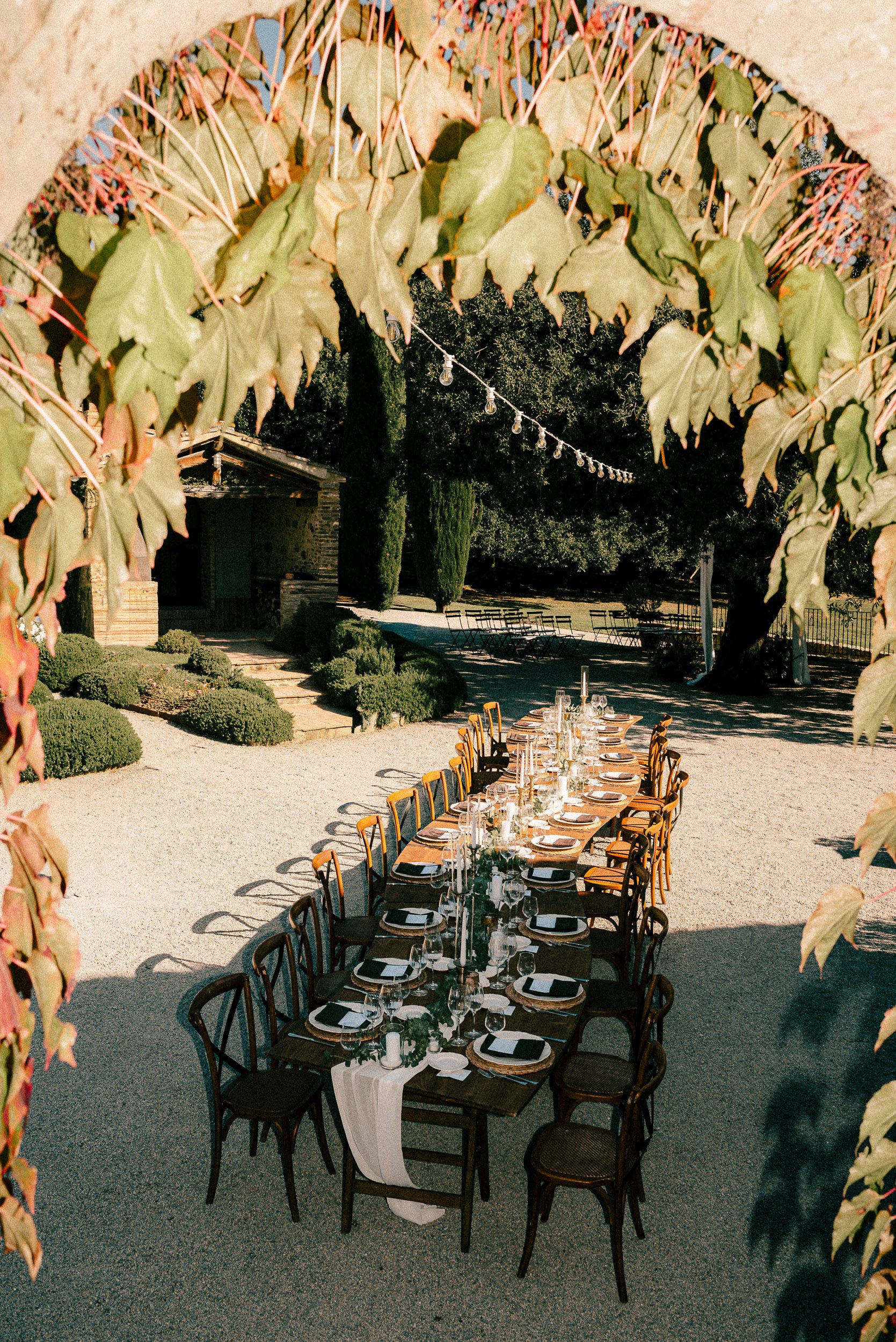 Outdoor dining setup with multiple tables and chairs arranged under leafy canopy in dappled sunlight.