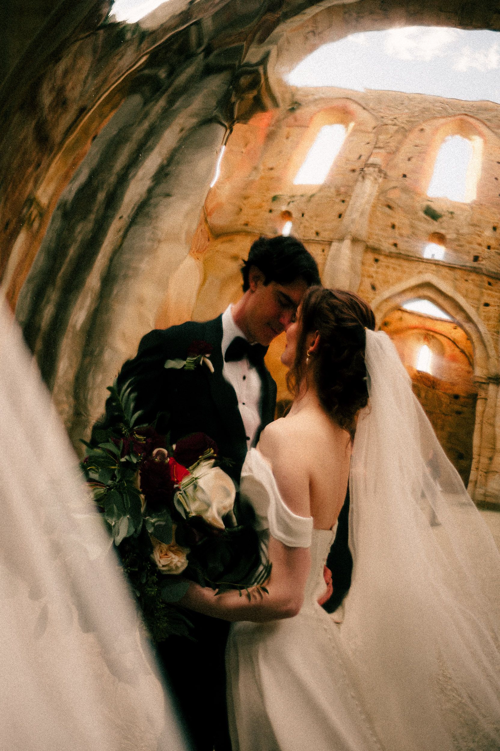 A romantic wedding couple shares intimate moments in a historic stone building with arched windows and natural light.