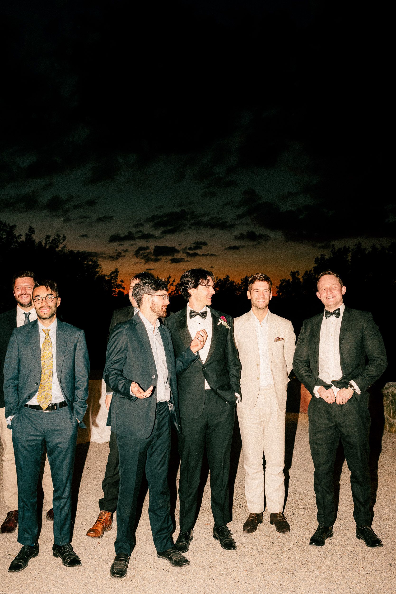 A line of groomsmen in formal suits and tuxedos stand together at dusk against a dramatic sunset sky.