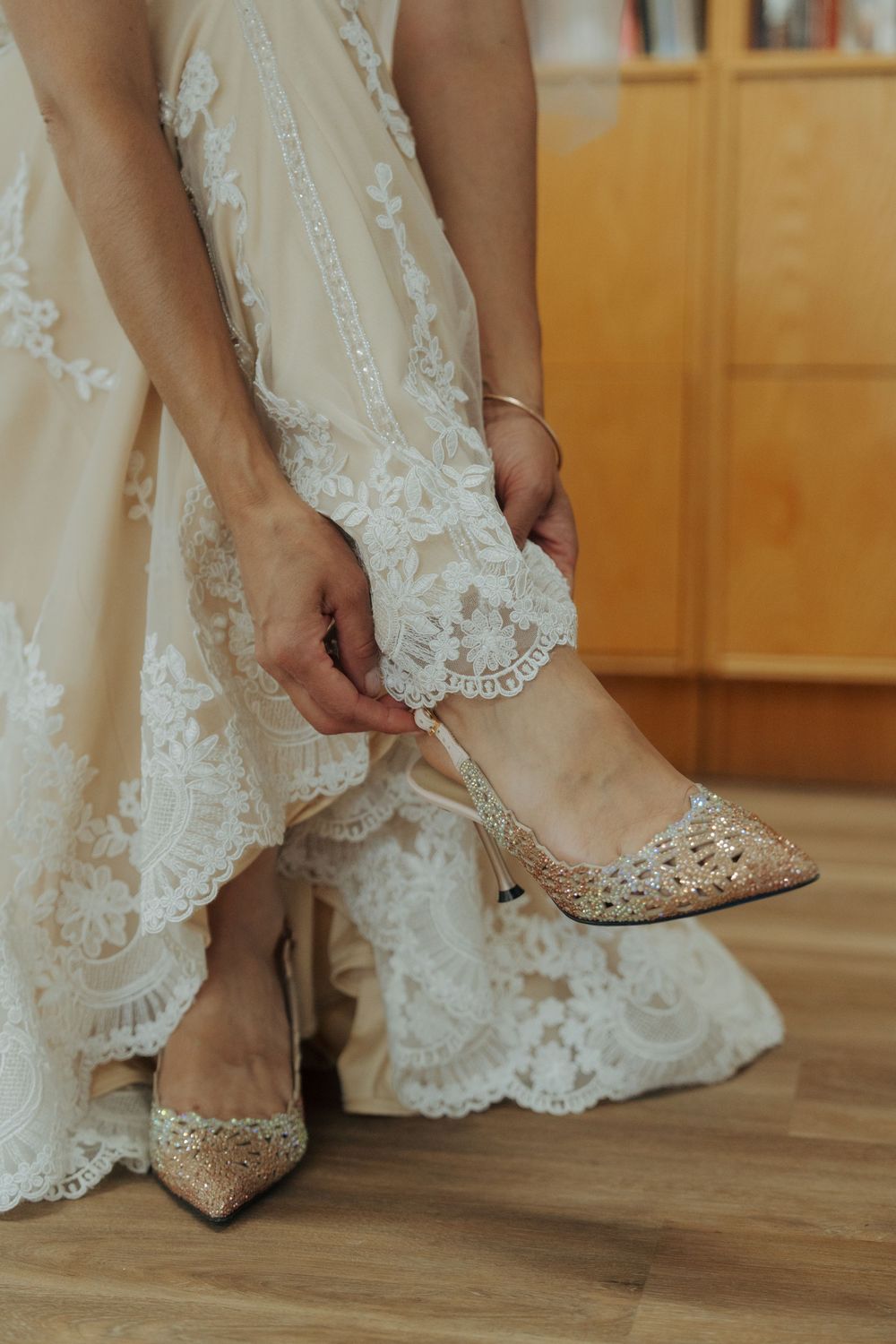 Bride putting on sparkly pointed-toe wedding shoes with delicate lace dress hem visible on wooden floor.