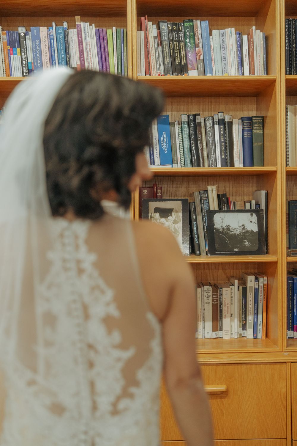 A bride in a lace-detailed wedding dress with a veil stands in front of wooden bookshelves filled with books.