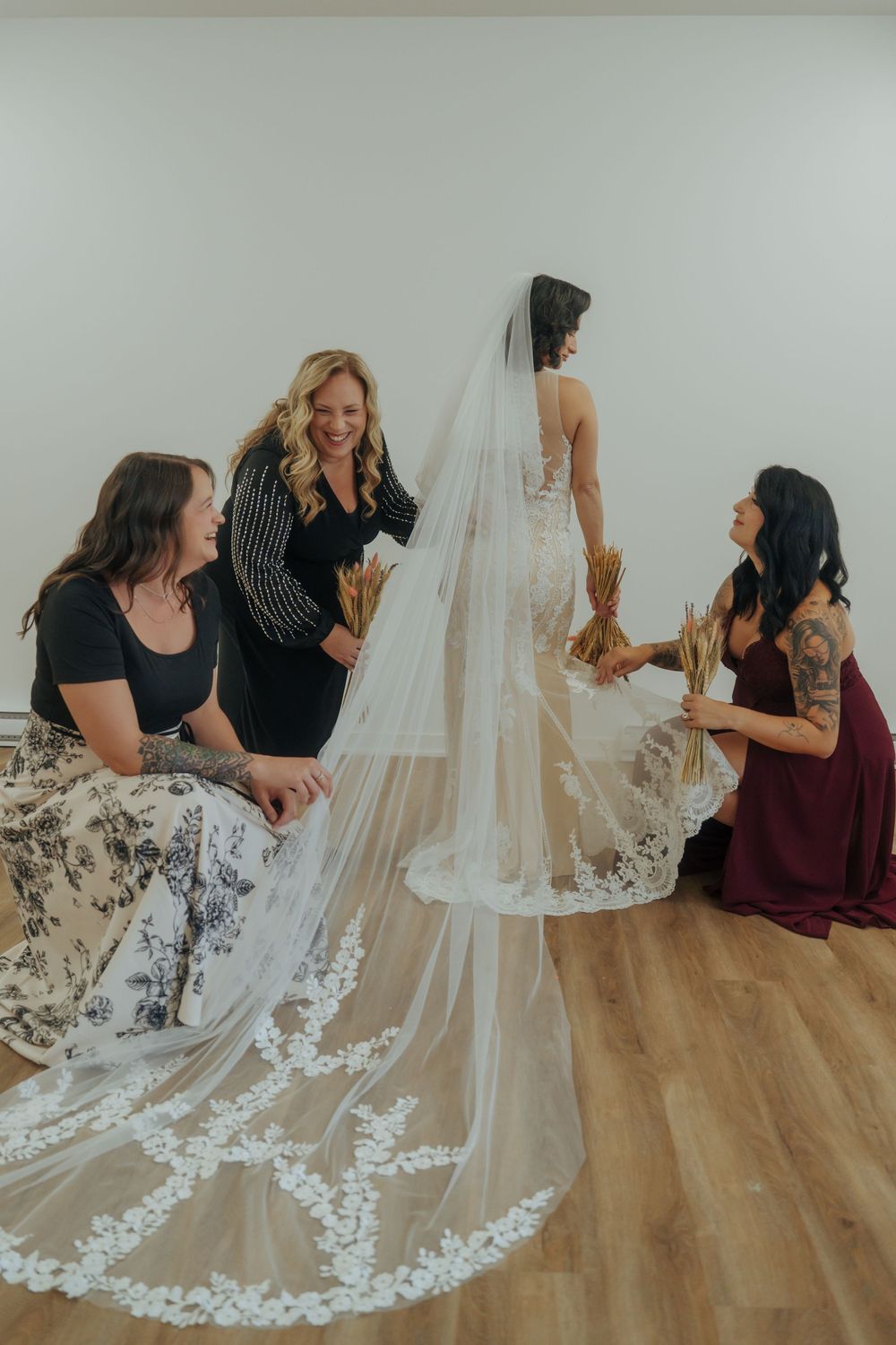 A wedding veil with intricate lace trim is being arranged on a hardwood floor by several people.