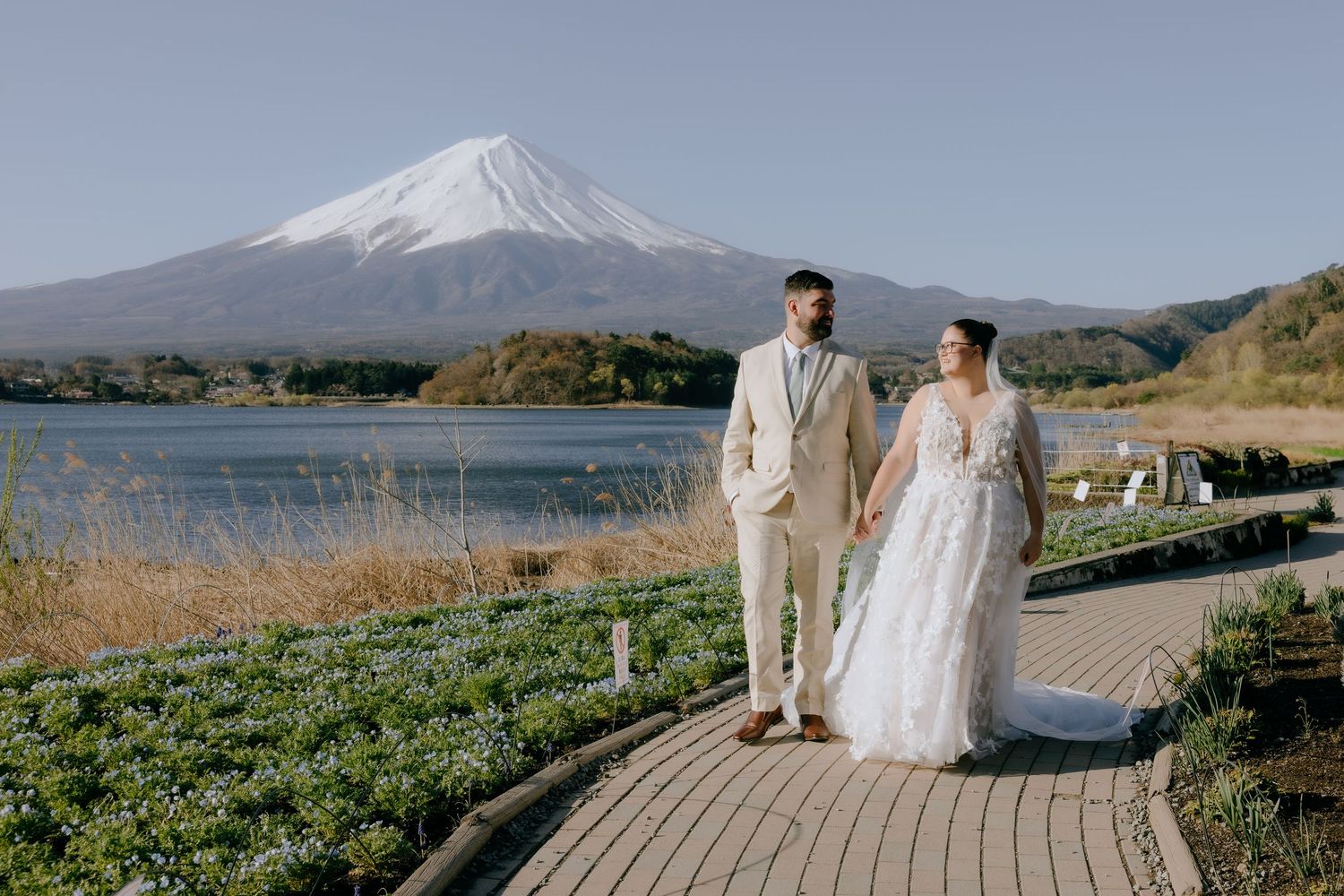A couple in wedding attire walks on a wooden pathway with Mount Fuji and Lake Kawaguchiko in the background.