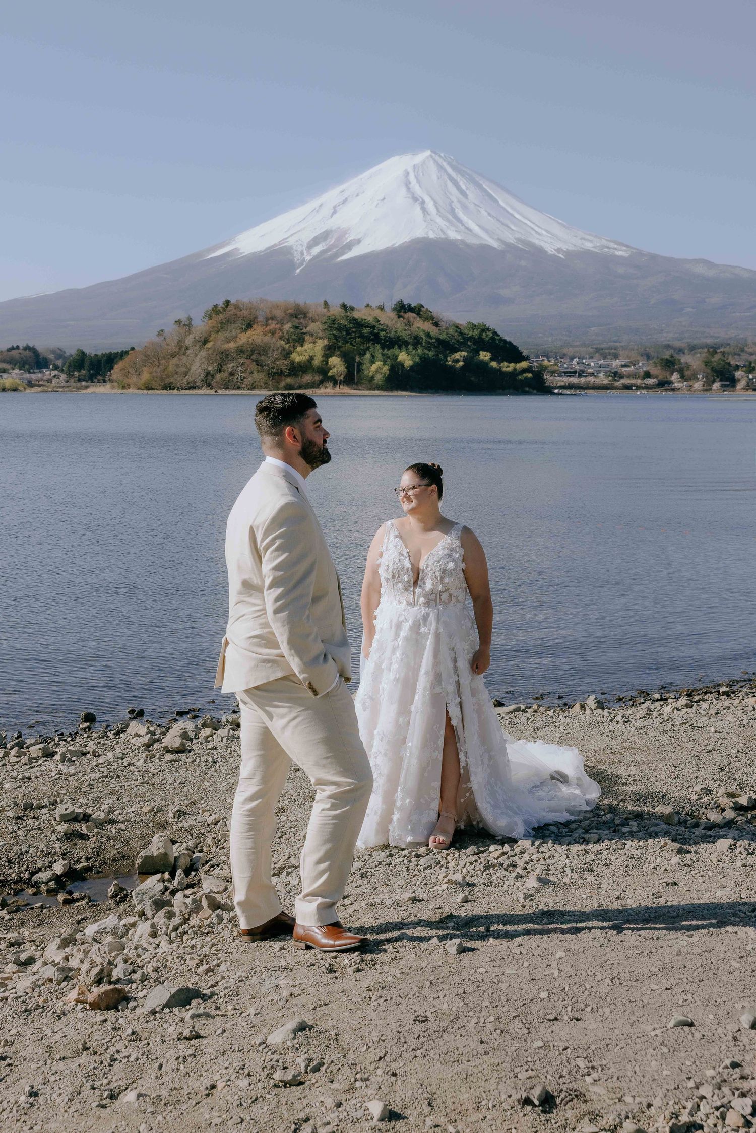 A couple sits together by Lake Kawaguchiko with majestic Mount Fuji rising in the background on a clear day.