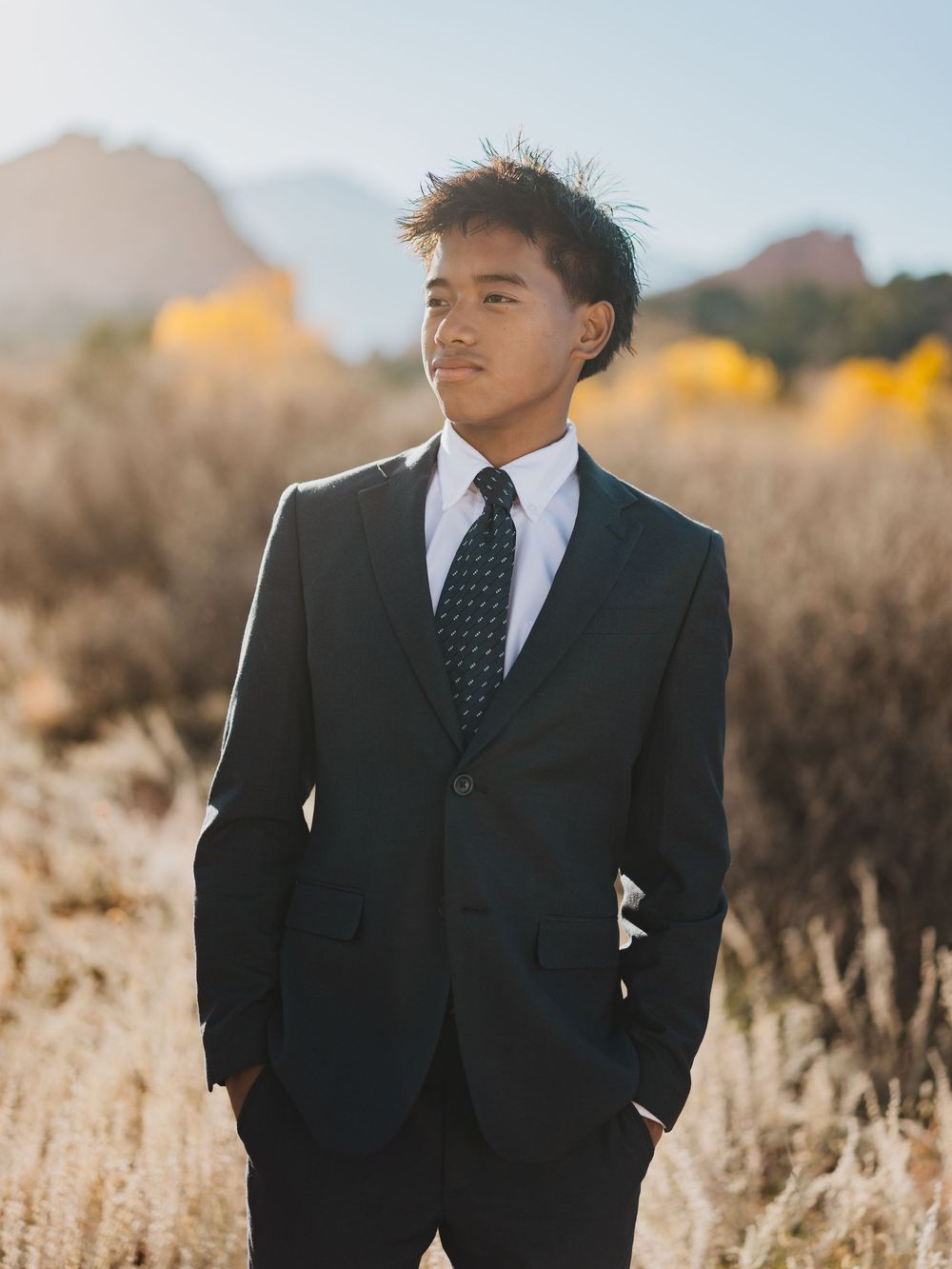 Young man in black suit and tie poses in desert landscape during golden hour sunset.