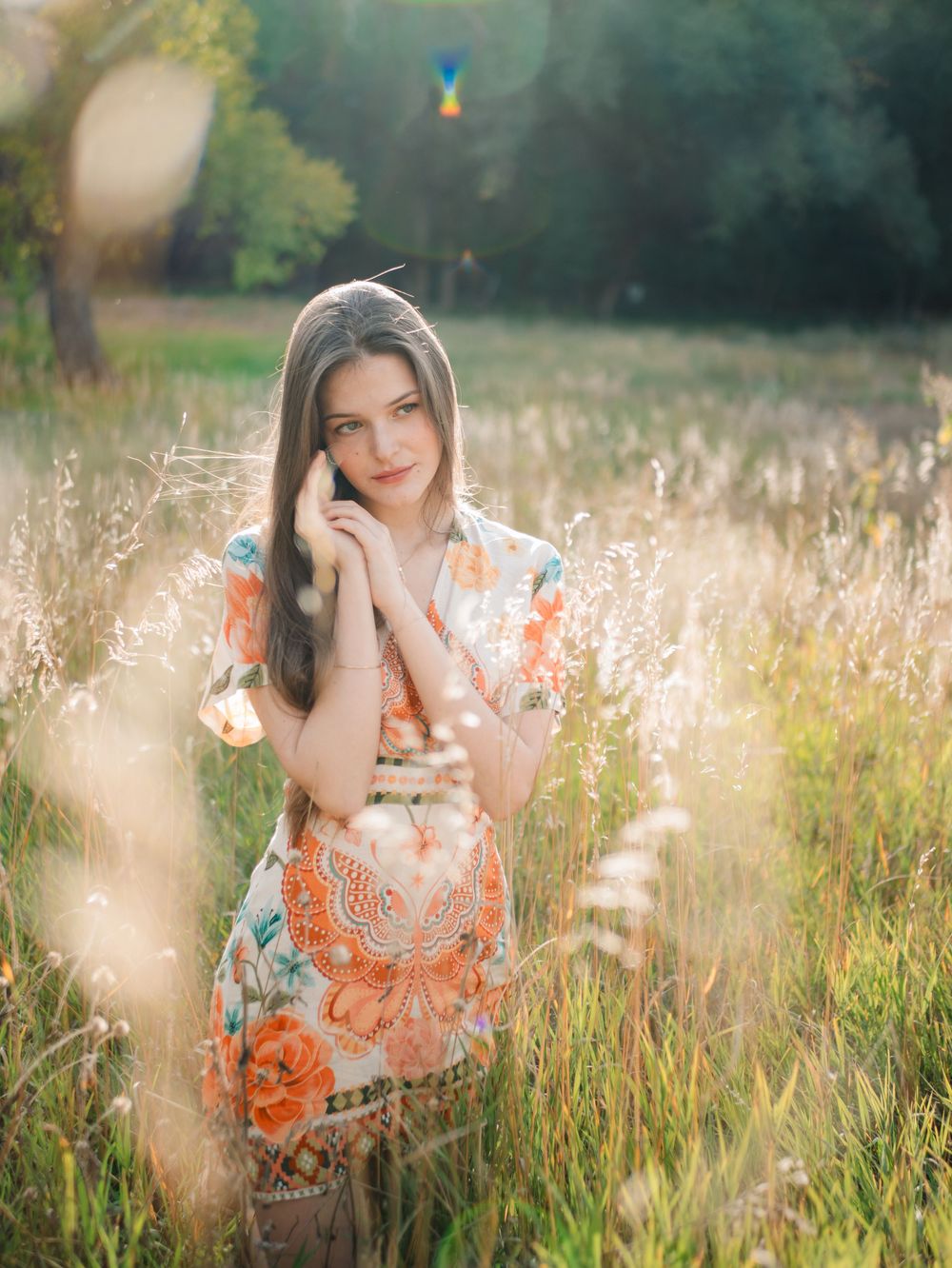 A woman in an orange floral dress poses playfully in a sunlit meadow with tall grass and wildflowers.