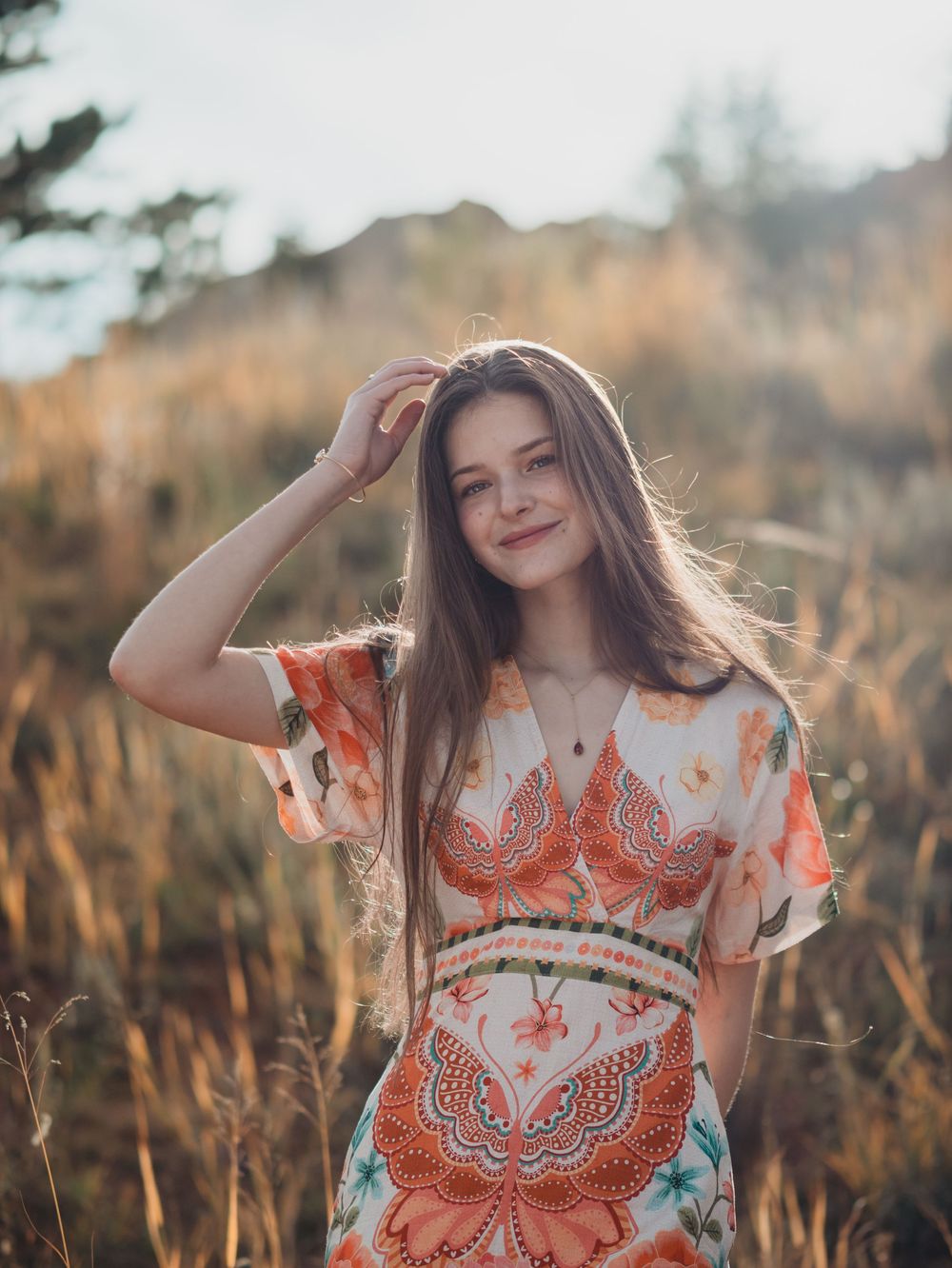 A person in a white and orange patterned dress poses gracefully in a sunlit field of tall golden grass at sunset.