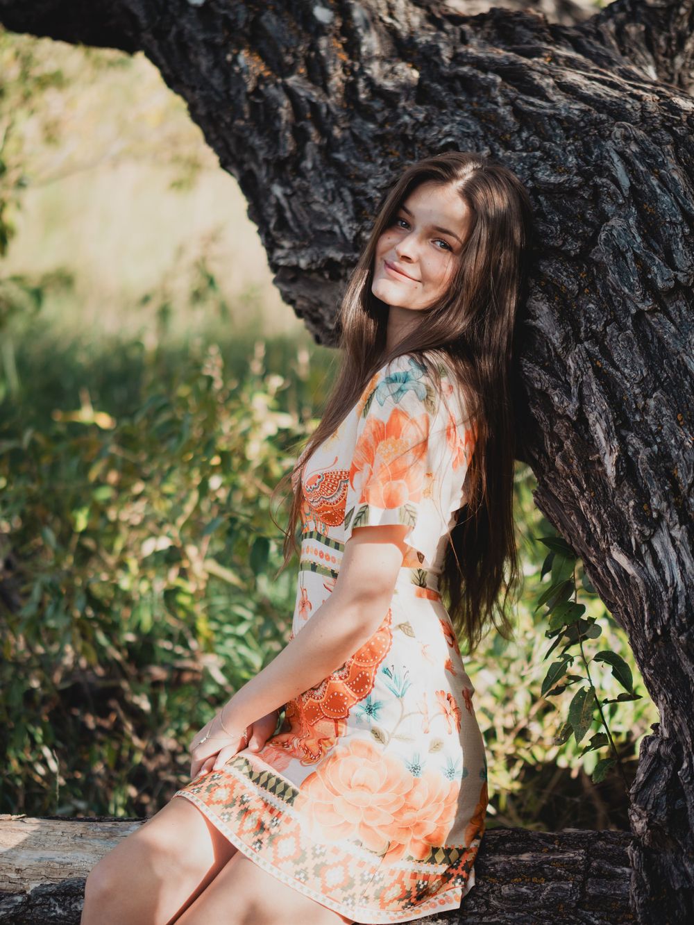 A person in a floral orange and white summer dress poses against a dark tree trunk in a sunlit outdoor setting.