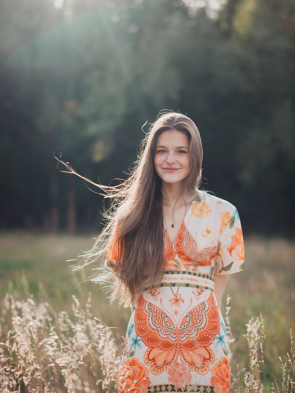 Person in orange and white butterfly print dress poses in sunlit meadow with tall grass and natural lighting.