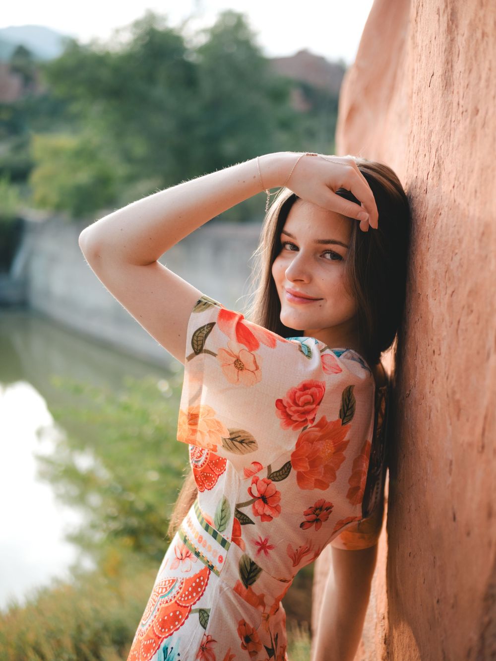 A person in a floral dress with orange roses leans against a peach-colored wall outdoors on a sunny day.