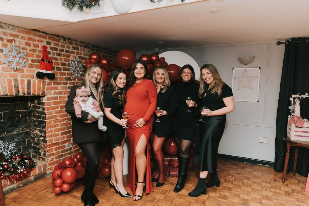 Group of friends celebrating at a holiday party with red balloons and brick wall decor.