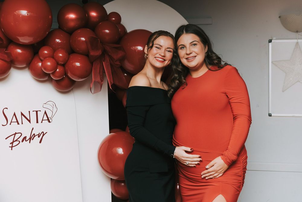 Two people pose together at a holiday party with red balloon decorations and a 'Santa Baby' sign on the wall.