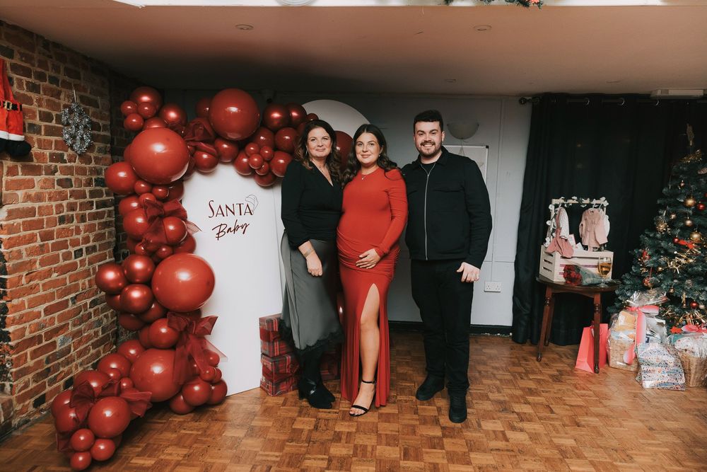 Red balloon arch decorates a brick wall interior during a festive celebration with people gathered around.