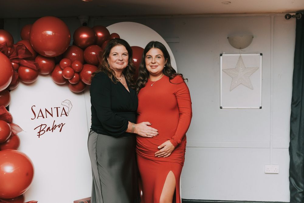 Christmas maternity baby shower photoshoot with red balloons and festive decorations against white wall.