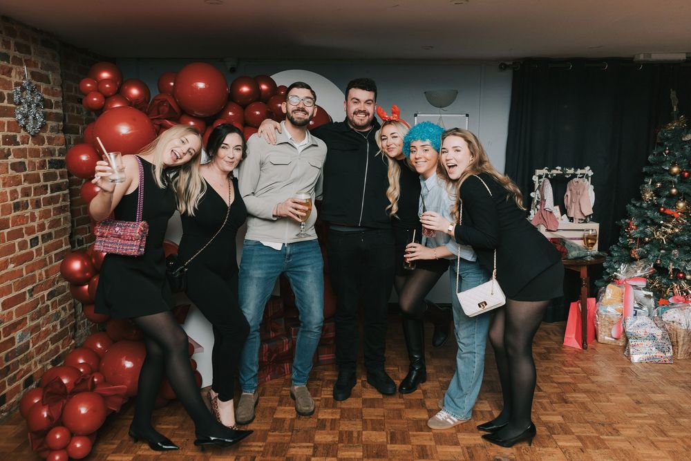 A group of people celebrate at a festive holiday baby shower party with red decorations and brick walls.