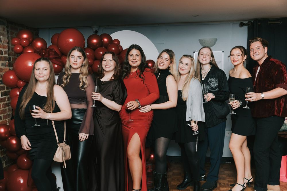 A group of people celebrating at a baby shower party with red balloons and black attire in a dimly lit venue.