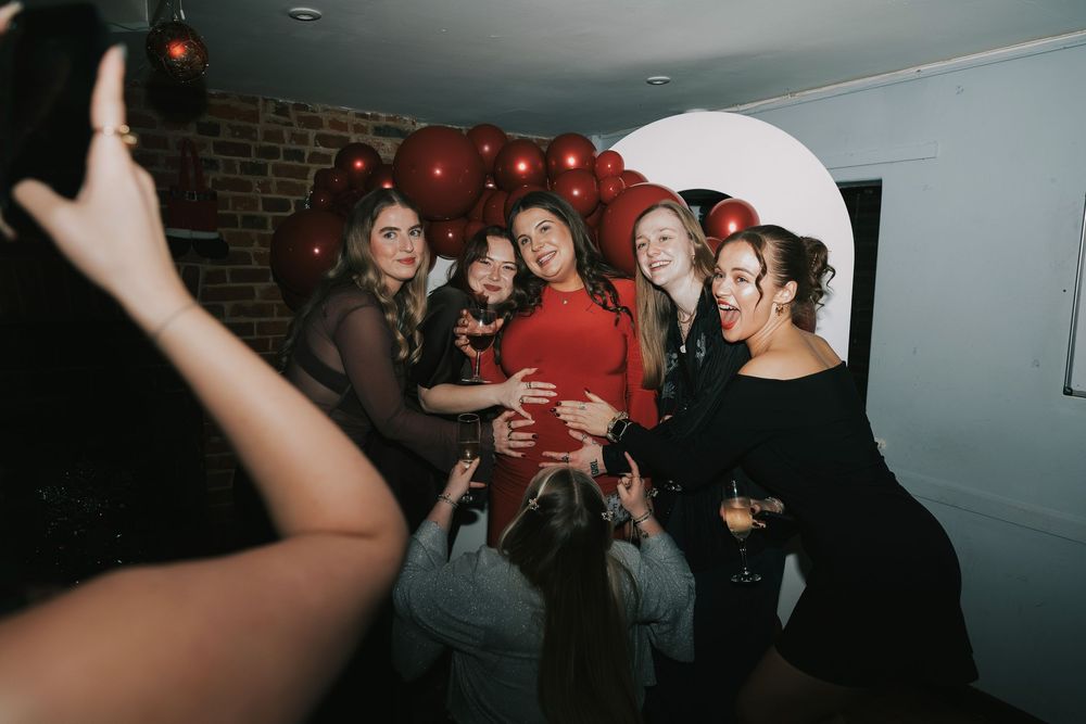 Partygoers gathered in a dimly lit venue around mum to be with red balloon decorations against brick walls.