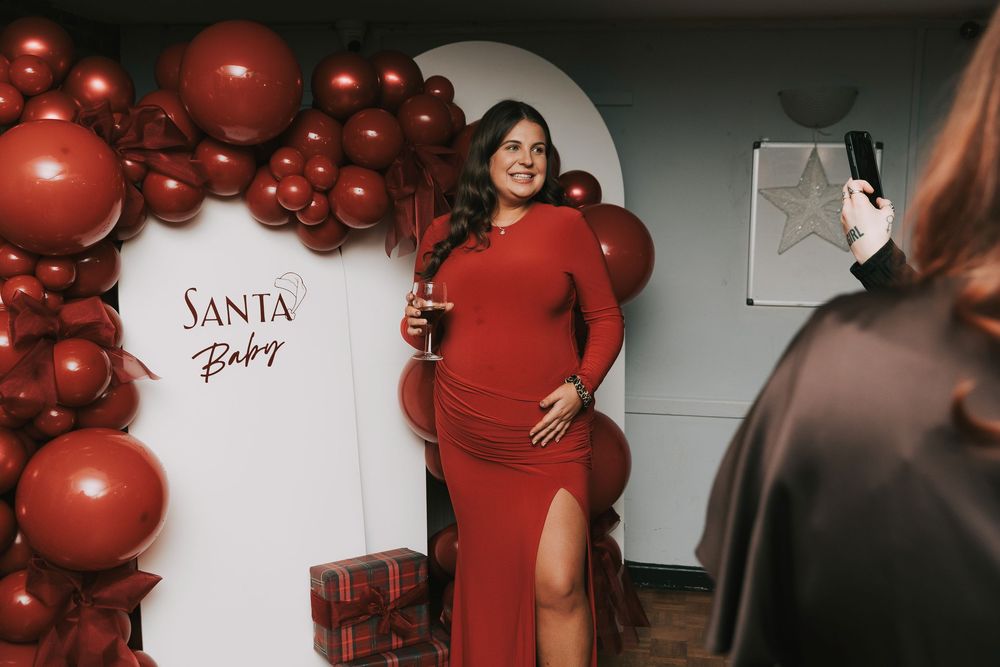 Holiday themed baby shower photo session with red balloon arch and Santa Baby sign on white backdrop.