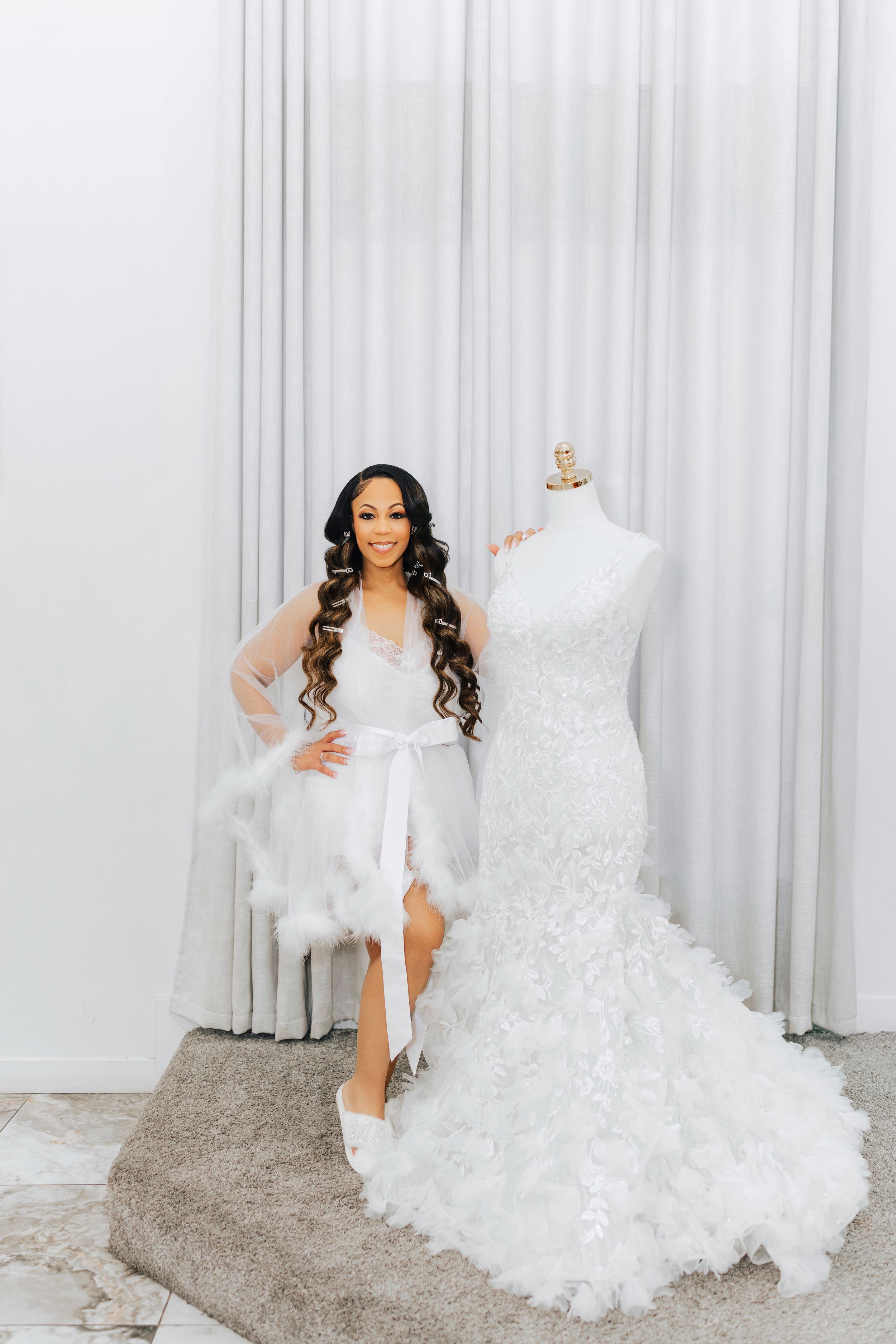 A woman in a white robe poses next to a mermaid-style wedding dress on a mannequin against white curtains.