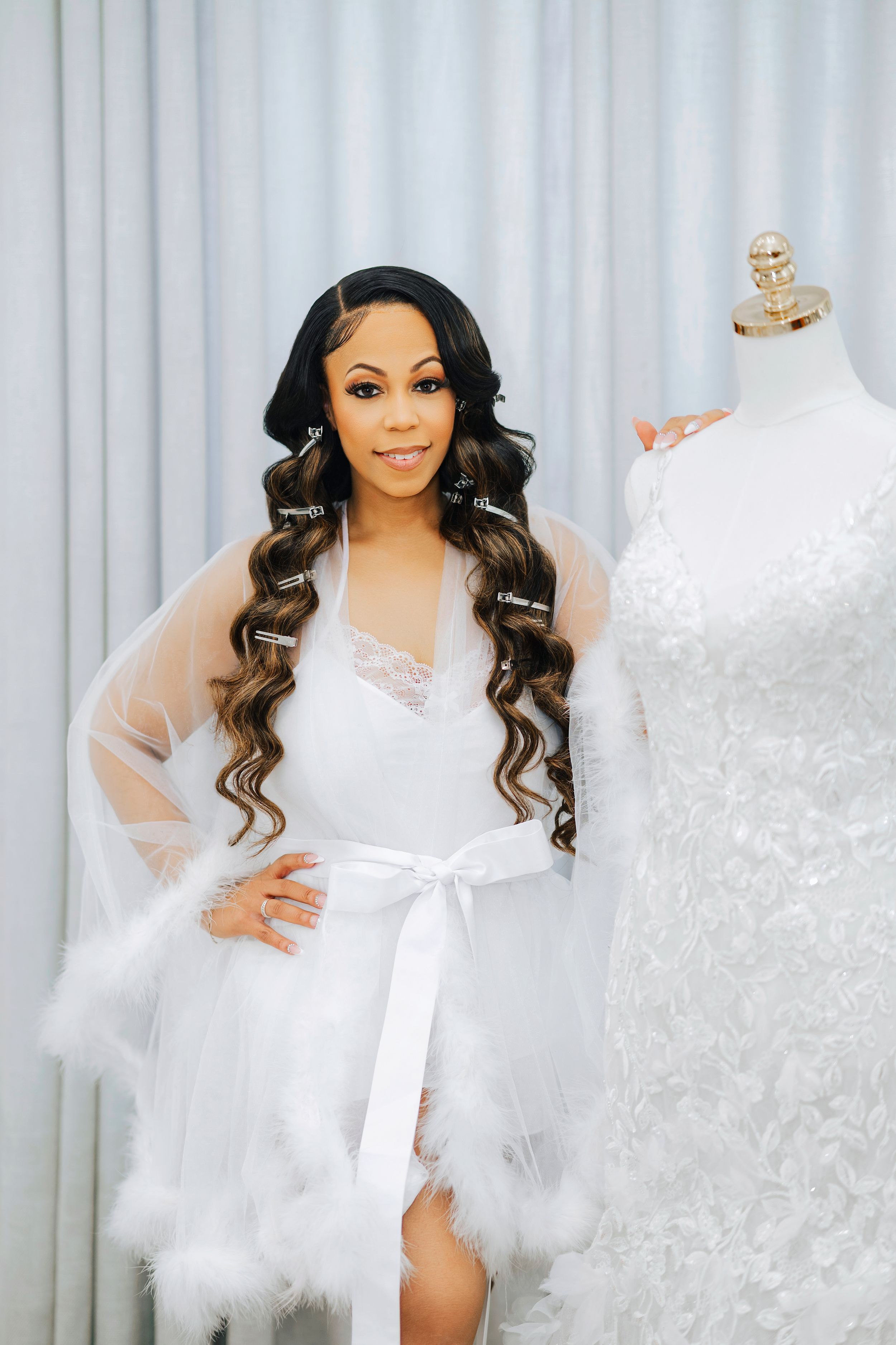 A bride in a white feather-trimmed robe poses next to a wedding dress on a mannequin in a bridal boutique.
