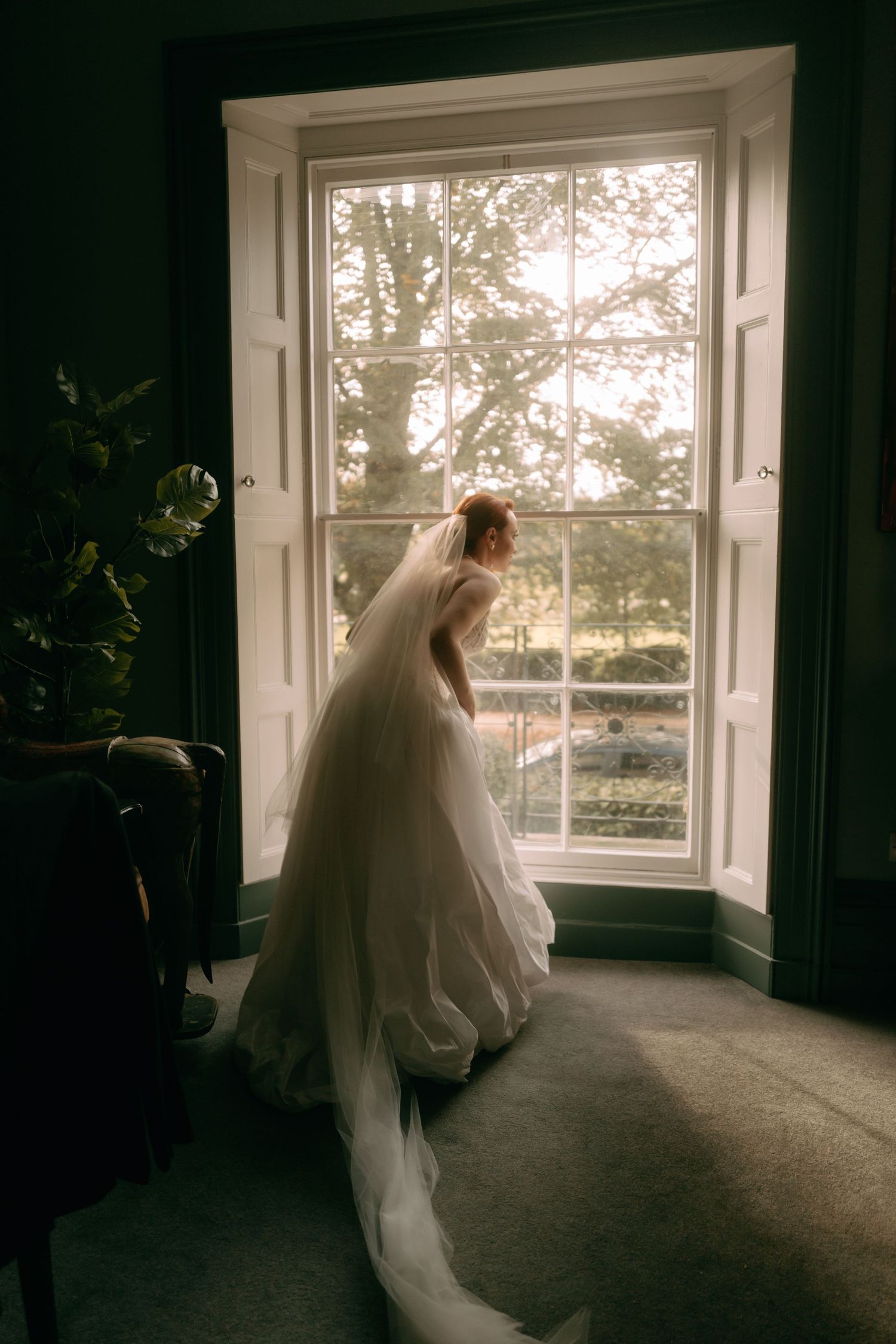 A bride in a flowing white wedding gown and veil stands silhouetted against a bright window in a dark room.