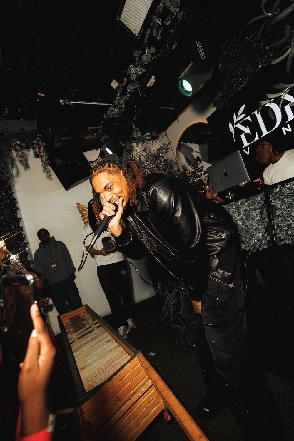 A performer sings passionately into a microphone at a dimly lit music venue with wooden flooring.