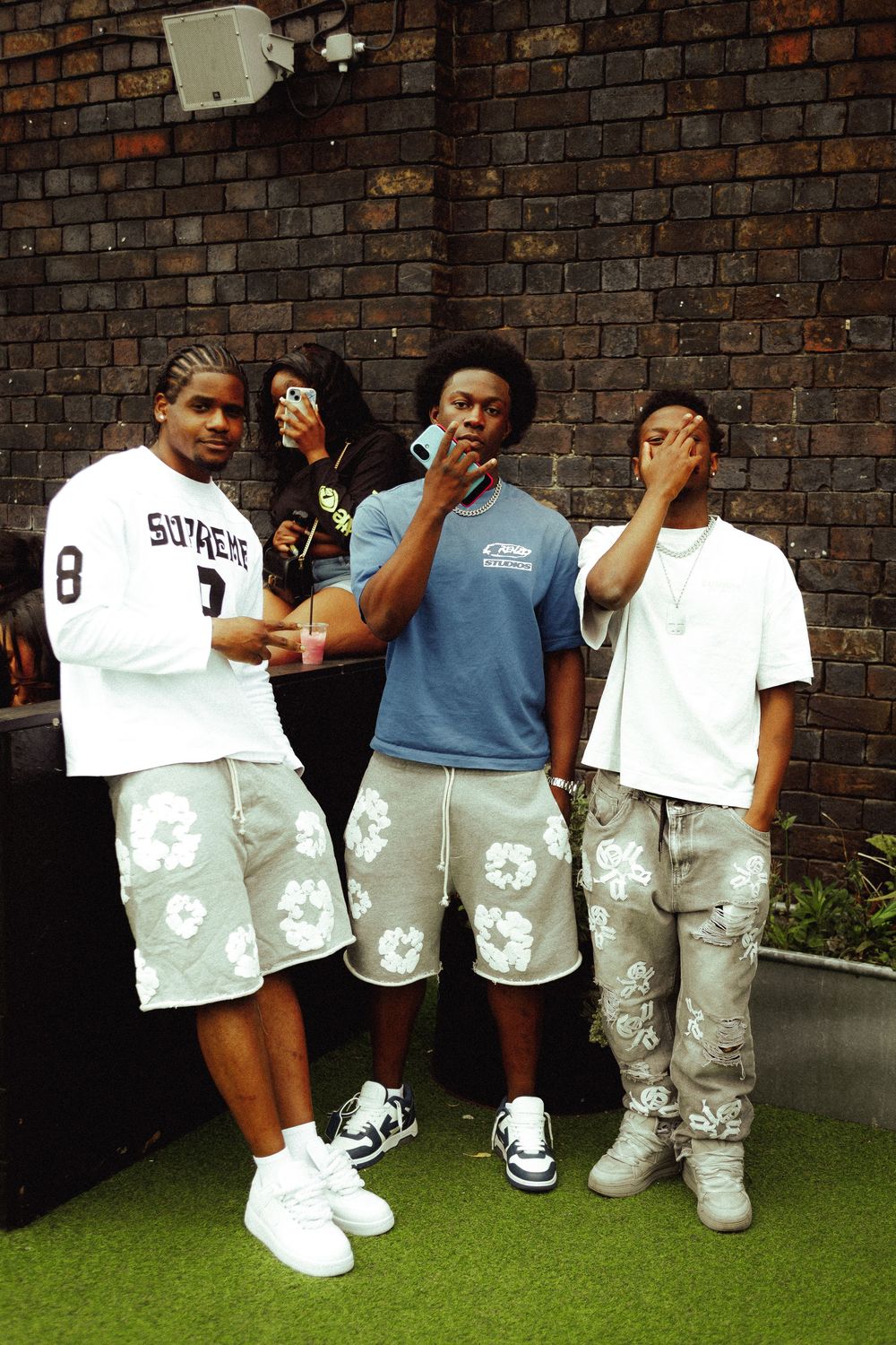 Four young men posing together outdoors against a brick wall, wearing casual streetwear including floral shorts and white sneakers.