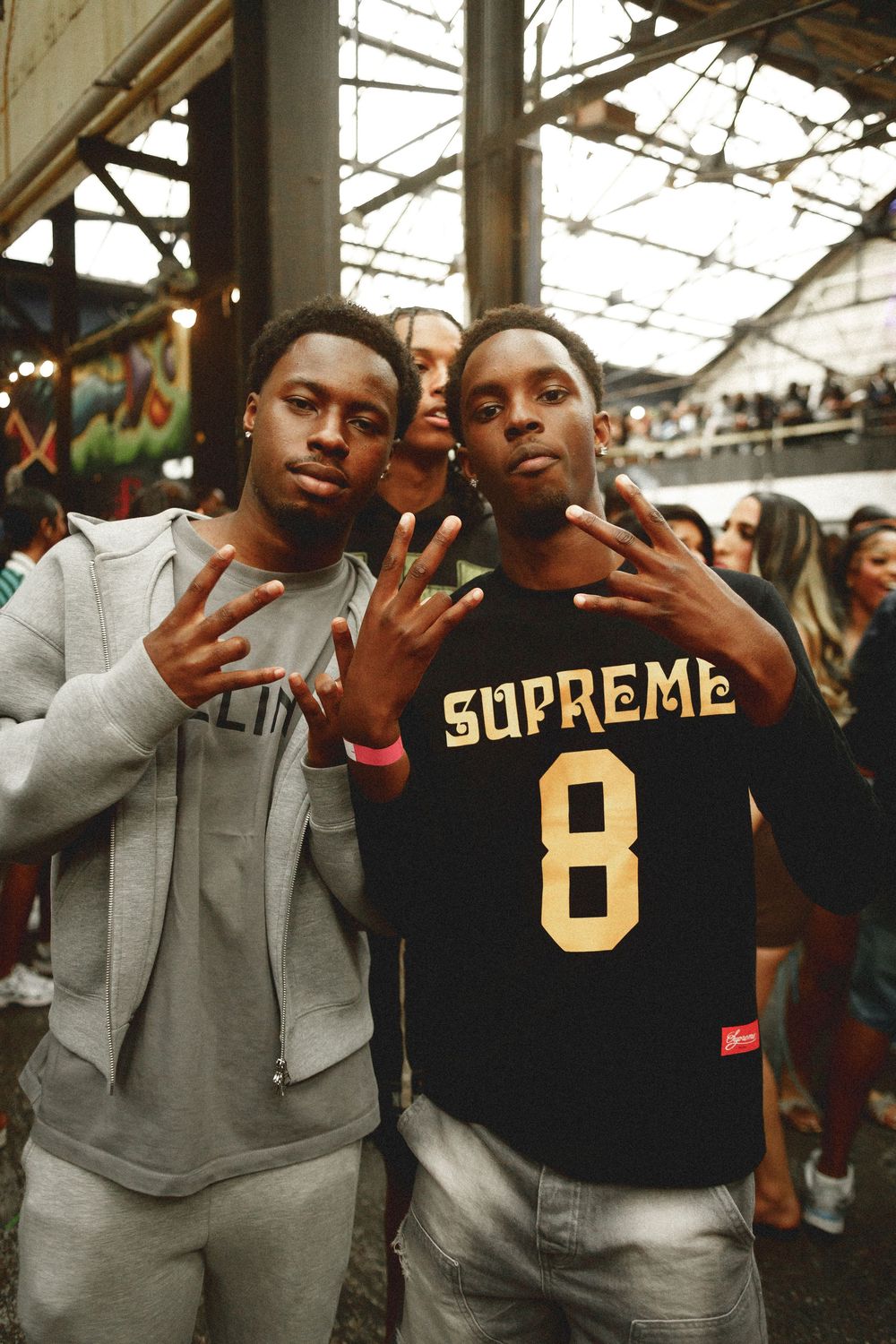 Two young men posing together flashing peace signs, one wearing a black Supreme jersey number 8 at an indoor event.
