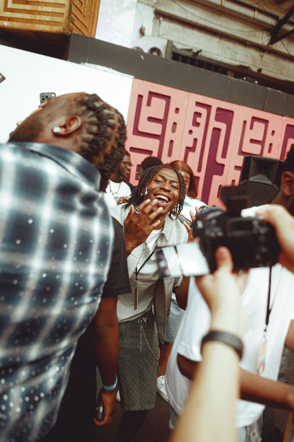 A joyful Black woman with dreadlocks laughs while being photographed at an indoor event with pink artwork backdrop.
