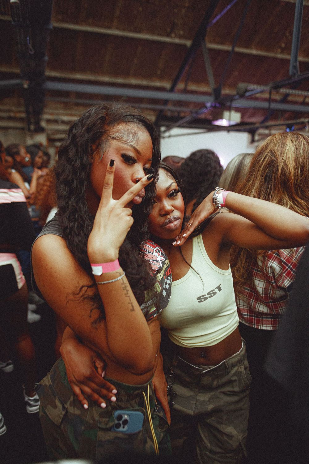 Two young women posing together at a party, flashing peace signs and smiling in stylish crop tops and camo pants.