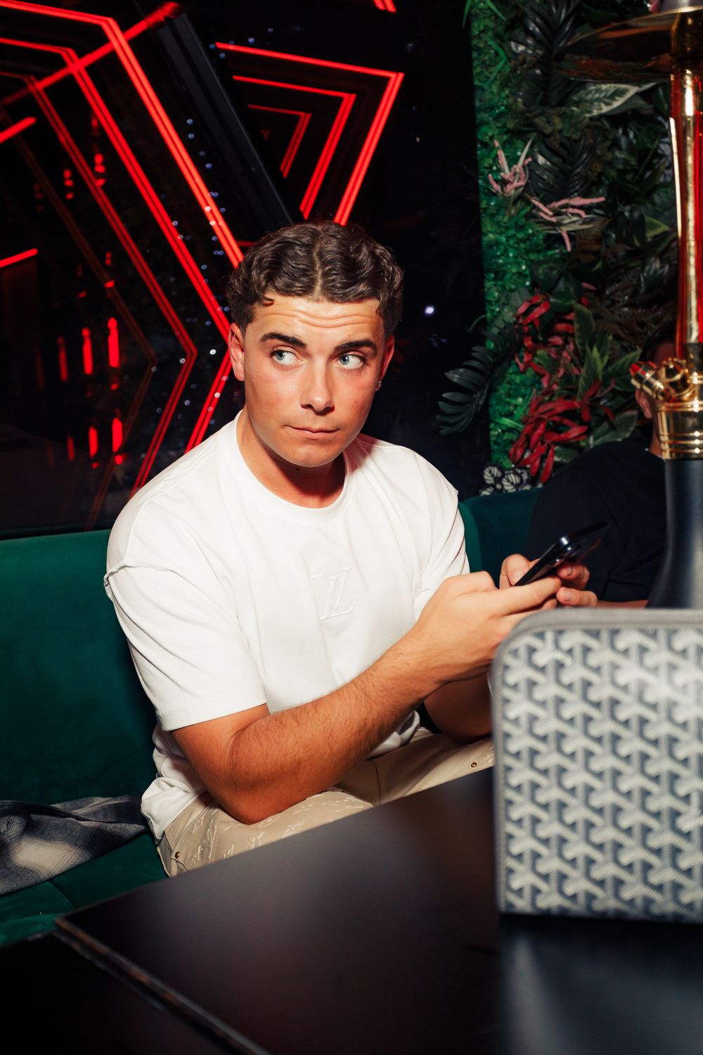 Person in white t-shirt sitting at table with neon red triangles and greenery in dark nightclub setting.