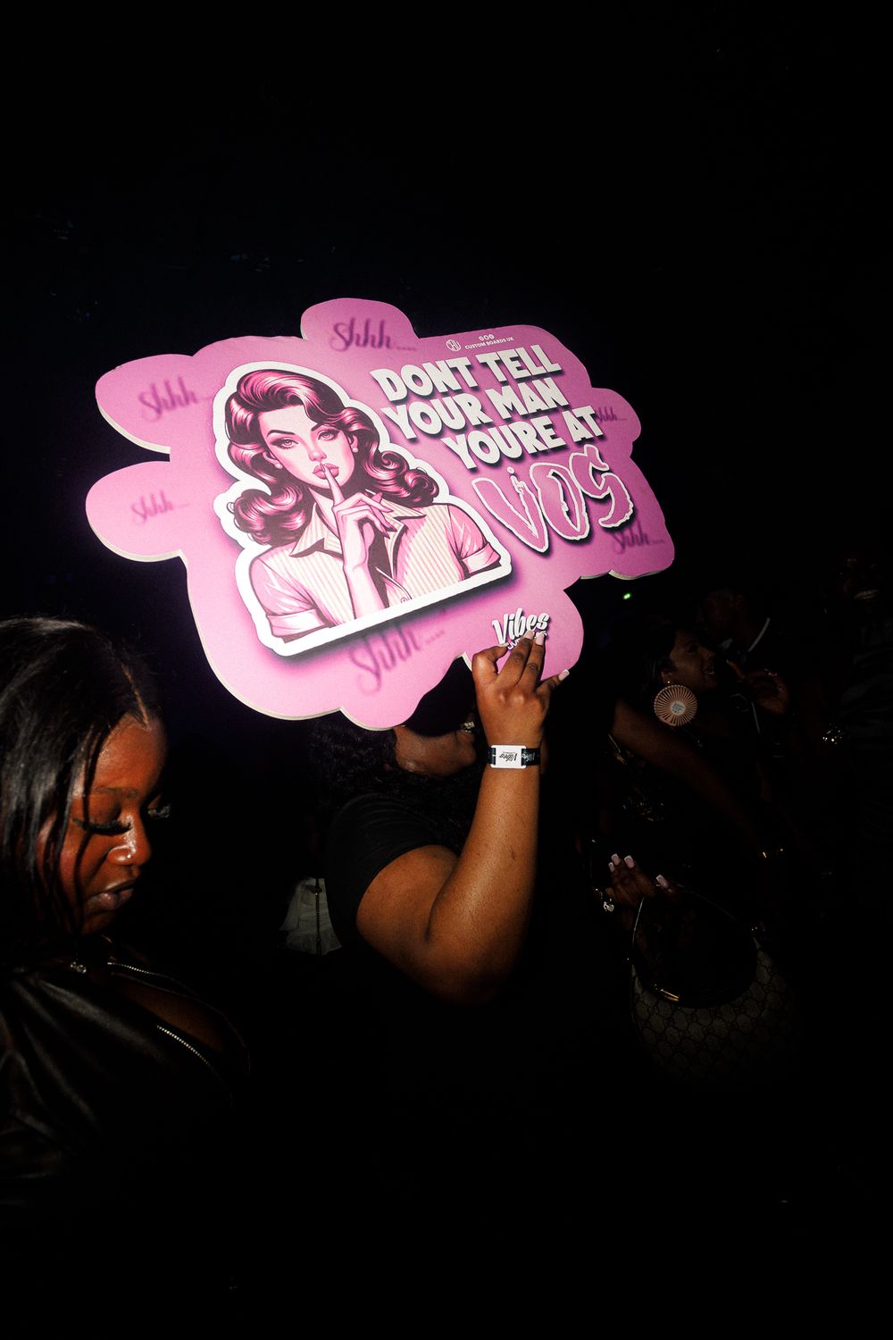 Pink promotional signs being held up at a dark concert venue creating a glowing effect against the black background.