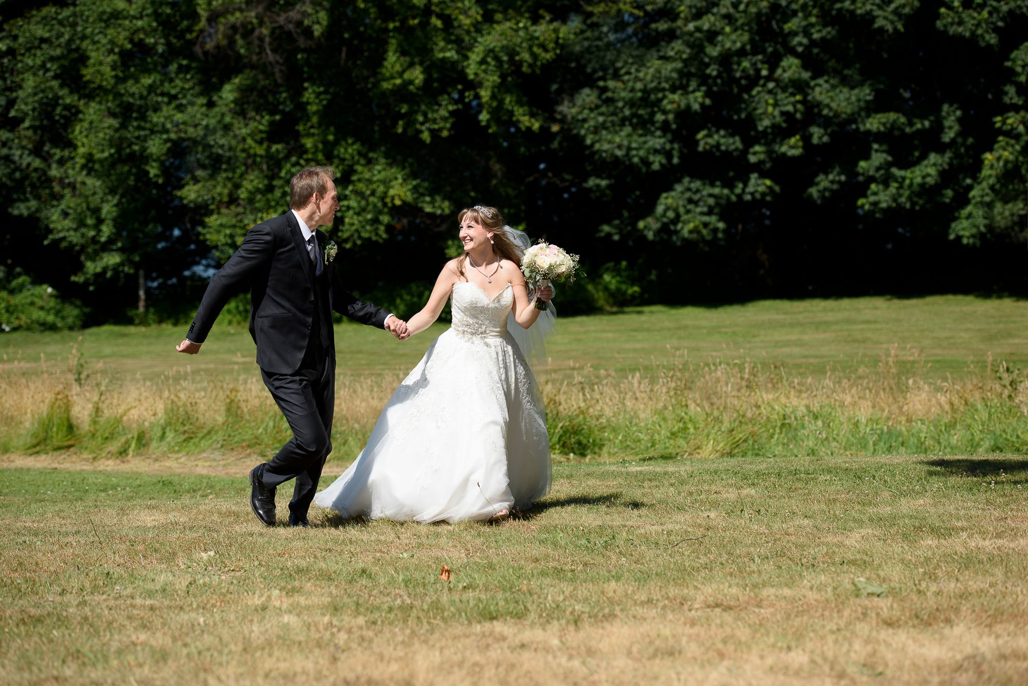 A couple in wedding attire playfully runs through a grassy field on their special day.