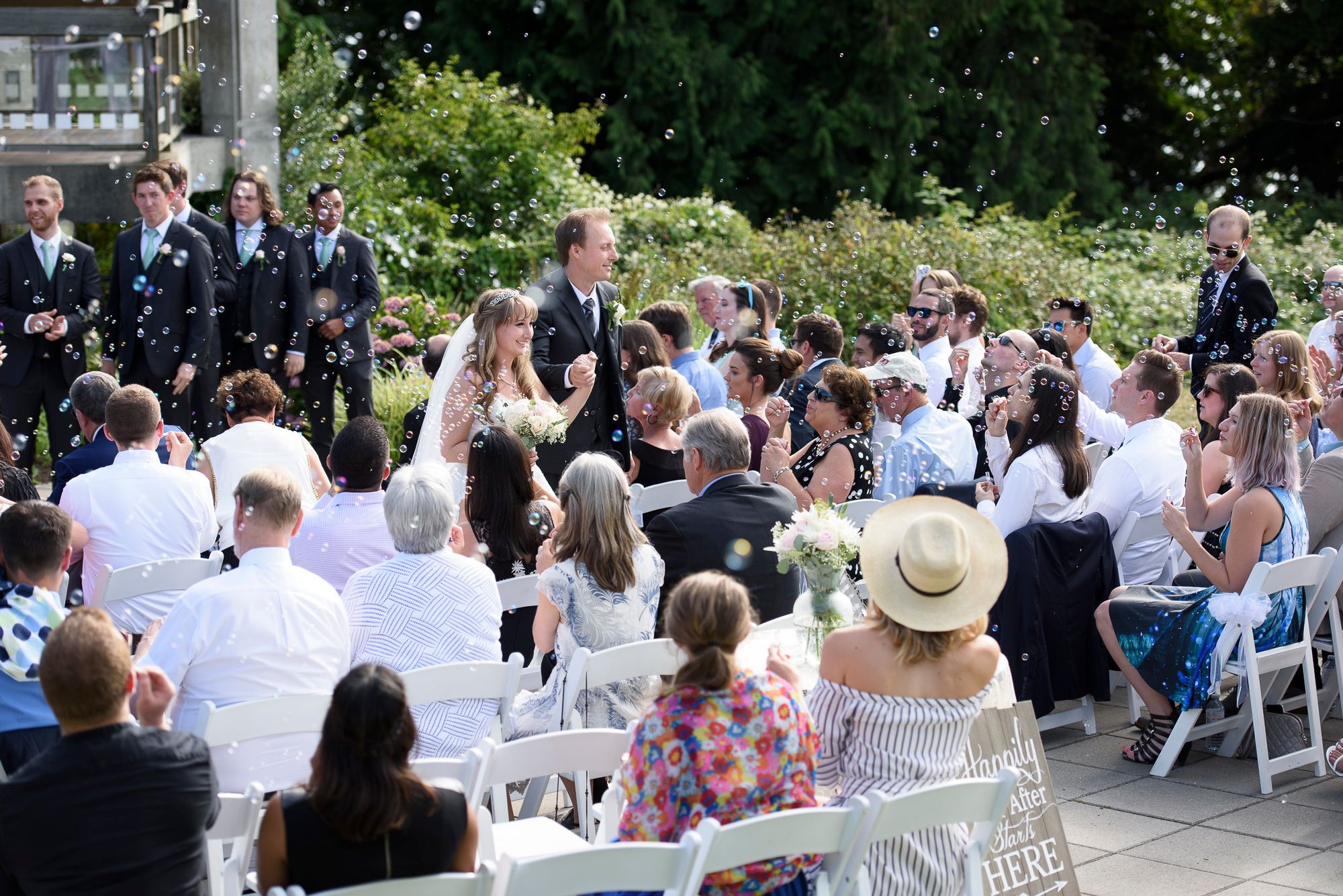 Outdoor wedding ceremony with guests seated in white chairs surrounded by lush greenery on a sunny day.