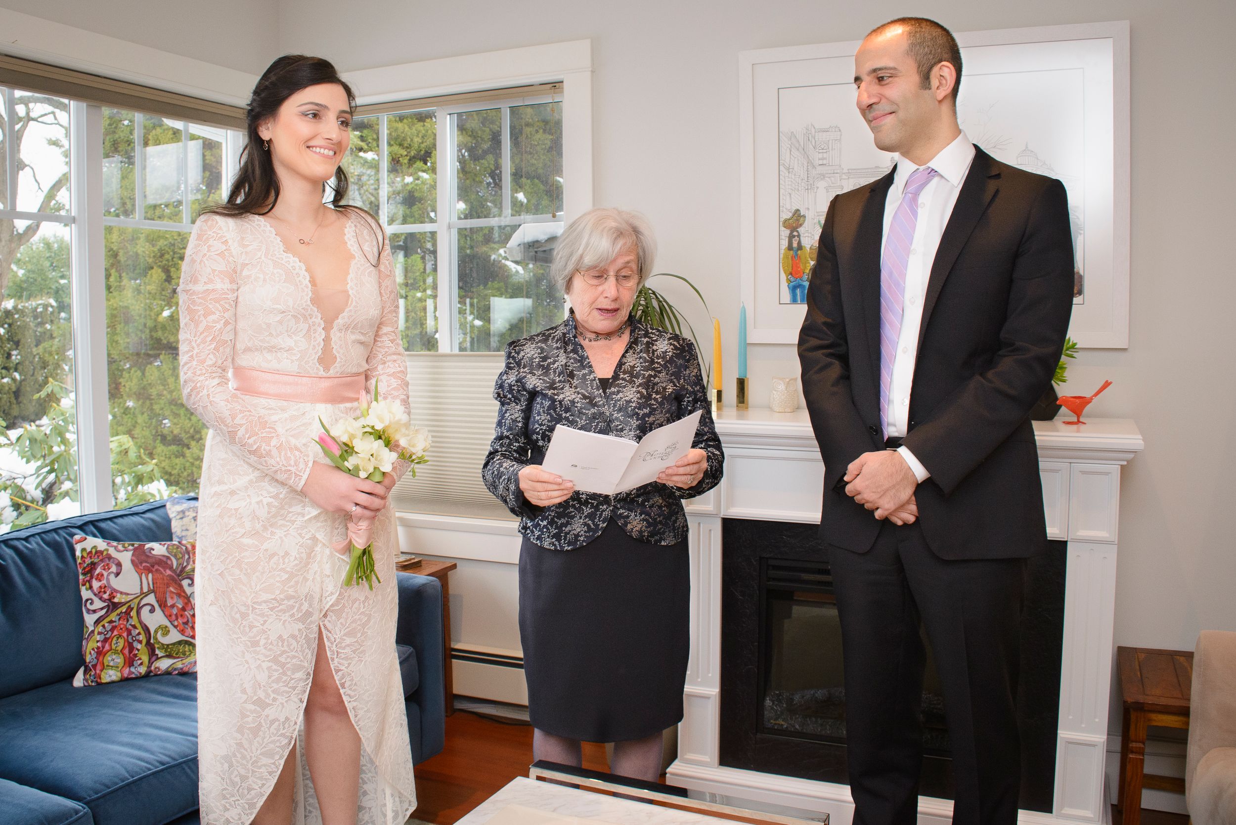 A small wedding ceremony takes place in a bright sunlit room with a blue couch and white fireplace.