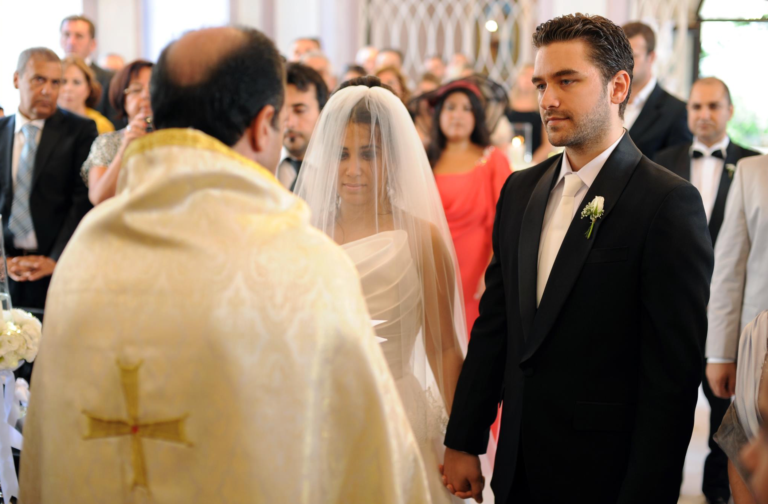 A wedding ceremony takes place in a bright church with people gathered and clergy in traditional robes.
