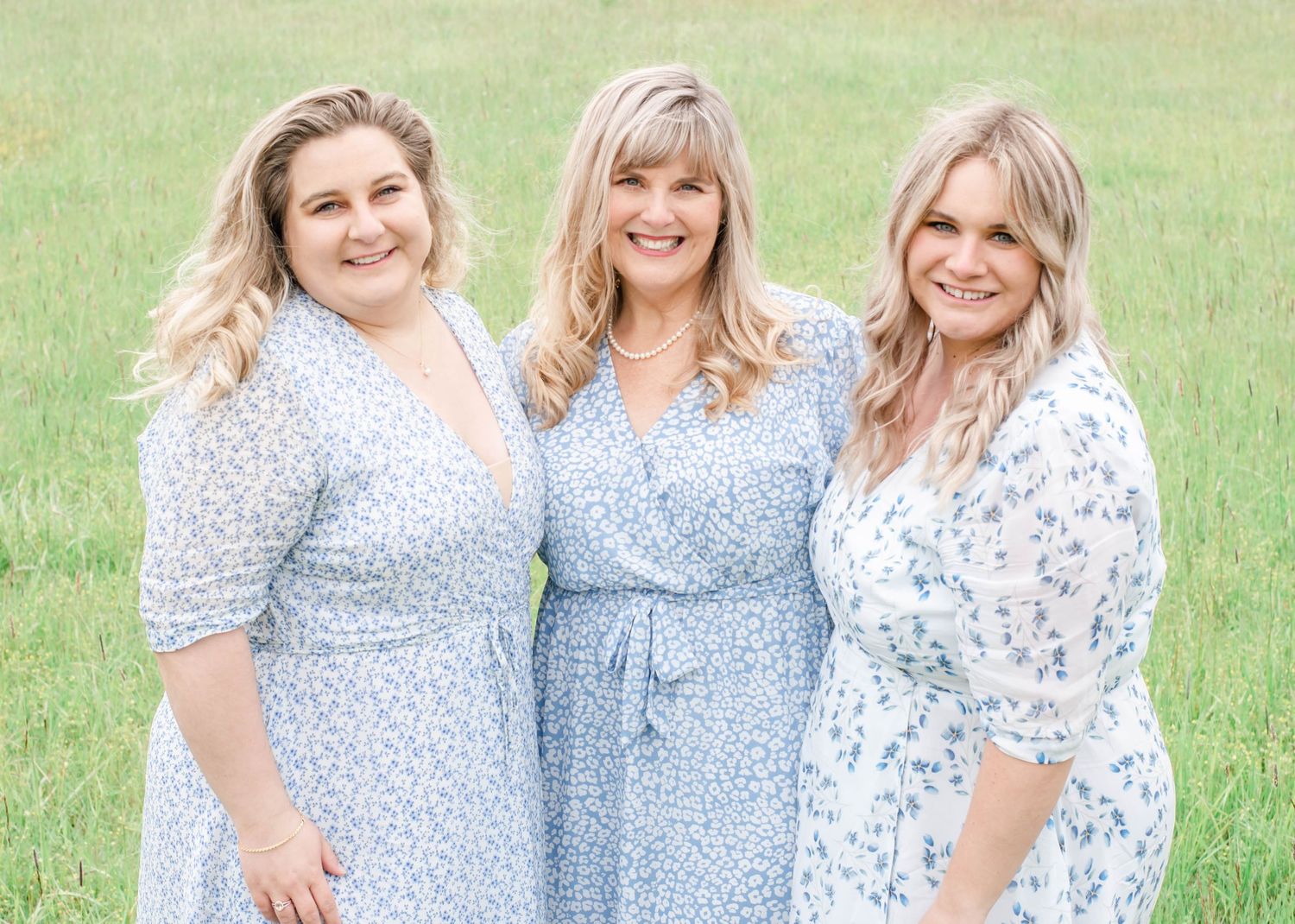 Three people stand in a grassy field holding a white flowing fabric in the air while wearing light colored clothing.