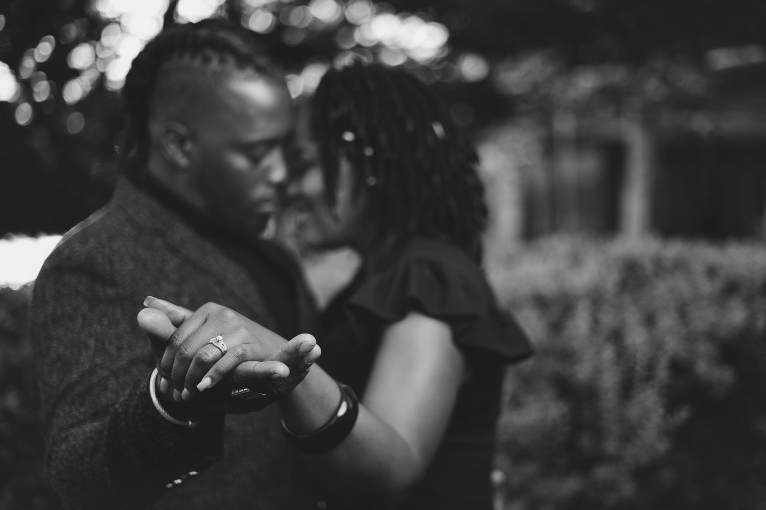 Black and white photo of a couple sharing an intimate moment in an outdoor setting with soft bokeh background.