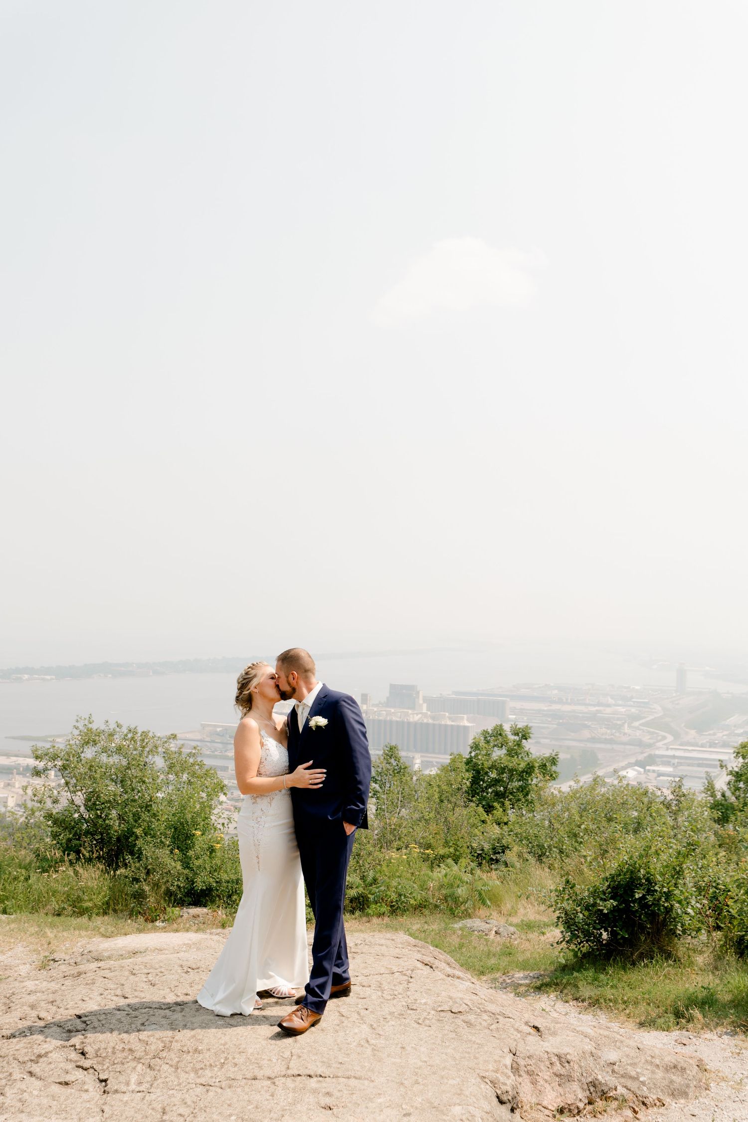 Bride and groom share a romantic kiss on a hilltop overlooking a city skyline on their wedding day.