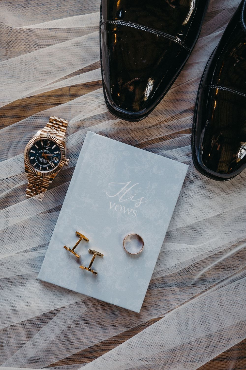 Gold cufflinks and watch arranged on gray paper with black shoes in the background.