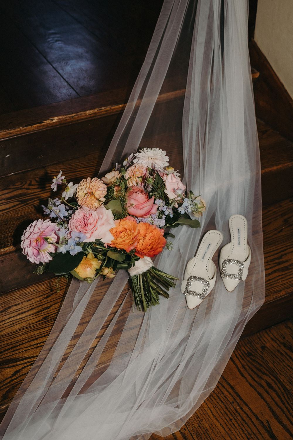 Bridal accessories including white shoes, veil, and colorful floral bouquet arranged on wooden floor.