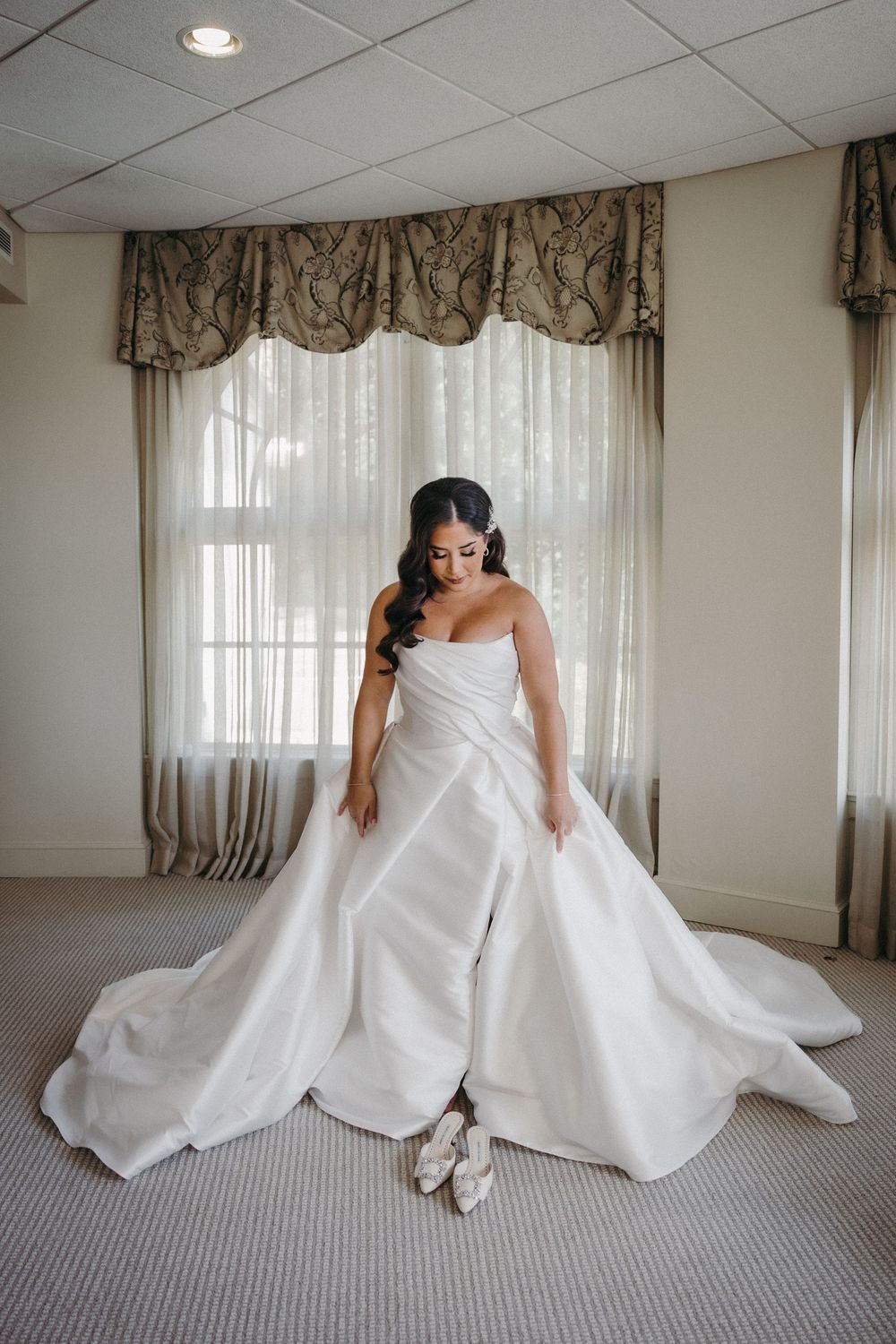 Bride sitting on carpet showing off her wedding dress in a sunlit room.