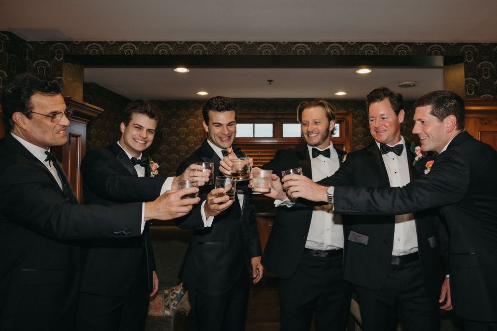 Groomsmen in black tuxedos toasting with drinks at a wedding celebration.