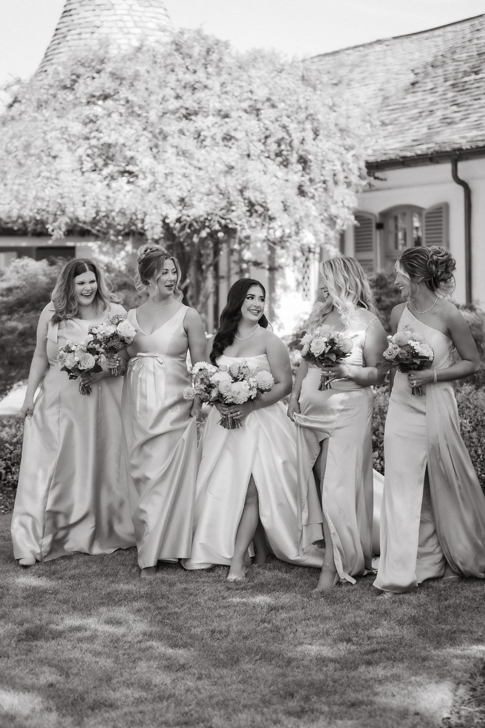 Bridesmaids in light colored dresses sitting together on grass holding bouquets.