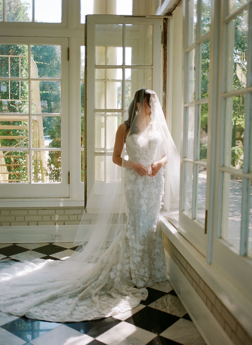 A stunning white wedding dress with a long train is showcased in a bright sunlit room with black and white checkered floors.