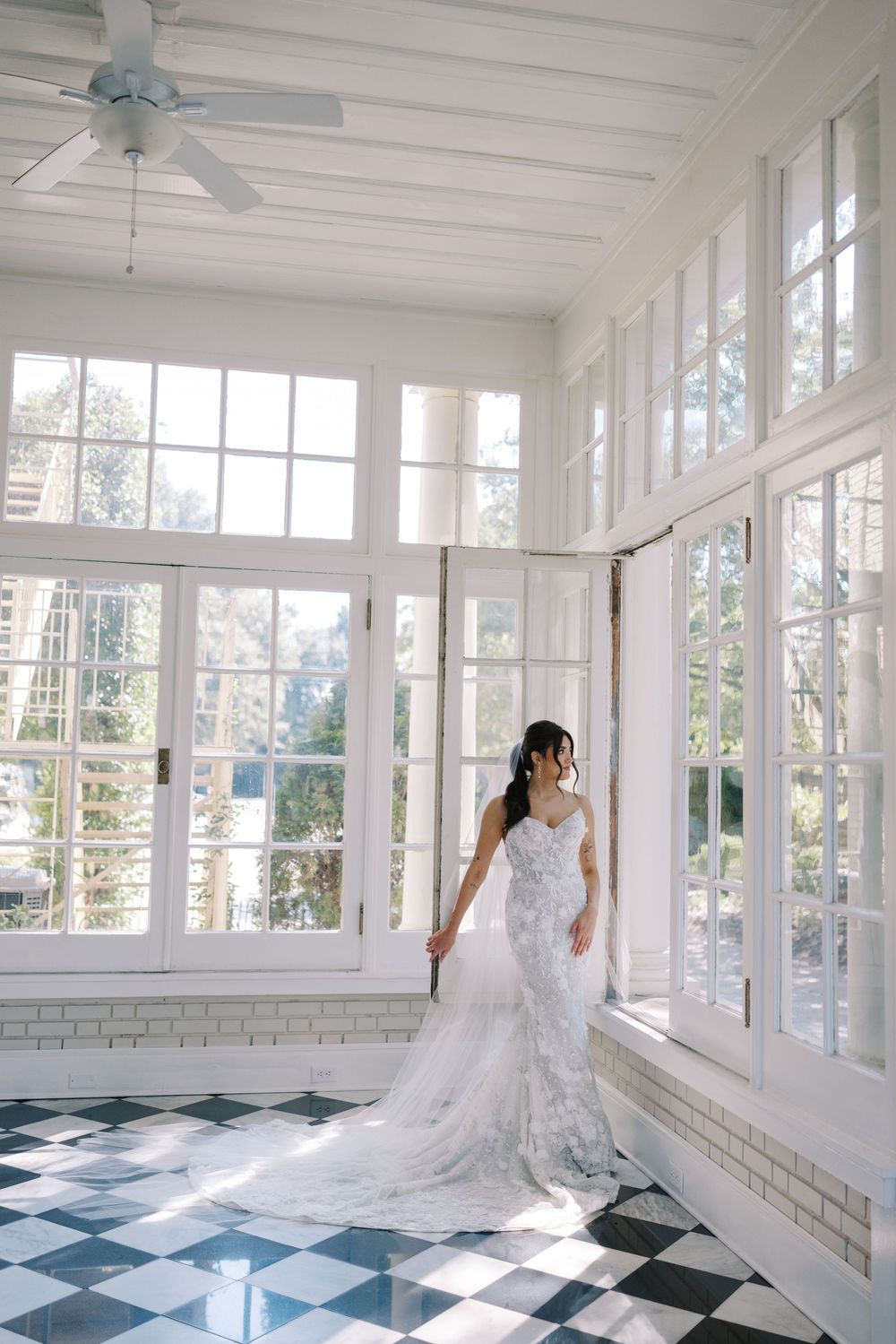 An elegant bridal gown flows across a black and white checkered floor in a sun-filled conservatory space.